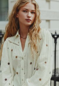 Light beige button-up shirt with embroidered geometric patterns in rust and blue. Features a relaxed fit and a delicate lace detail along the neckline.