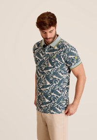 Polo shirt met een groen bladerenpatroon op een lichte achtergrond, voorzien van een gestreepte kraag en een klein logo op de mouw. Korte mouwen, klassieke pasvorm.