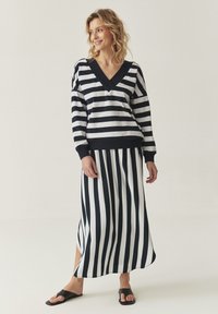 Maglione a righe navy e bianco con scollo a V, abbinato a una gonna lunga coordinata con righe verticali e uno spacco laterale. Sandali neri a scivolo.