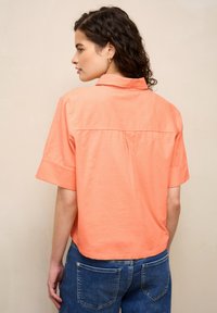Femme aux cheveux bouclés portant un t-shirt corail à manches courtes et un jean bleu, vue de dos sur un fond uni.