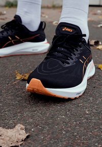 Black athletic shoe with white and orange details, textured upper, laces, and rubber sole. Ground surface includes scattered dry leaves.