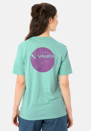 Donna vista da dietro che indossa una T-shirt verde menta con un design topografico circolare viola e il logo "VAUDE", abbinata a pantaloncini blu.
