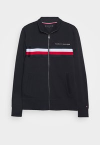 Černá zipová mikina má stojáček, boční kapsy a vodorovné pruhy v červené, bílé a modré barvě přes hrudník. Zobrazeno logo "TOMMY HILFIGER".