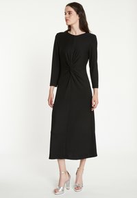 Robe midi noire avec un détail de nœud torsadé à la taille, manches longues et tissu lisse. Associée à des sandales à talons hauts argentées.