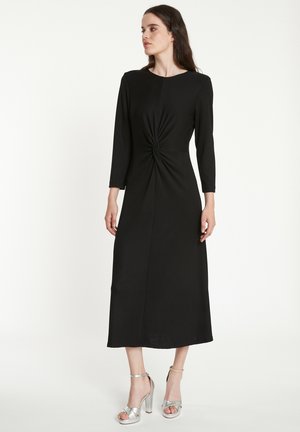 Robe midi noire avec un détail de nœud torsadé à la taille, manches longues et tissu lisse. Associée à des sandales à talons hauts argentées.