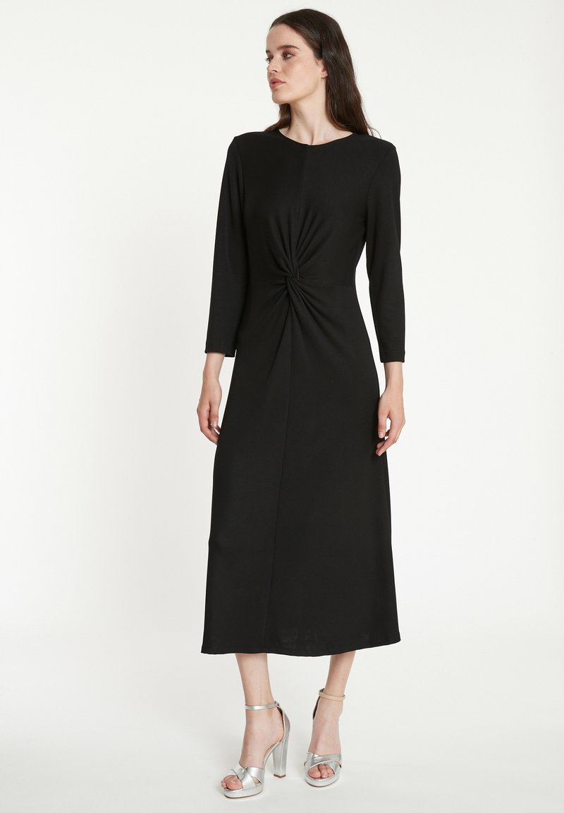 Robe midi noire avec un détail de nœud torsadé à la taille, manches longues et tissu lisse. Associée à des sandales à talons hauts argentées.