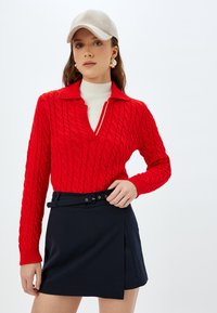 Czerwona sweter z dzianiny o głębokim dekolcie w kształcie litery V i beżową czapką. Noszony z granatową spódnicą z bocznym zapięciem. Teksturowany i dopasowany krój.
