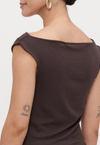 Sieviete valkā tumši off-shoulder topu, redzami zelta gredzena auskari, saules tetovējums un kursīva teksta tetovējums augšējā iekšējā rokā.