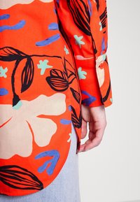 Chemise à fleurs rouges avec des contours noirs et des accents bleus et verts. Elle présente un ourlet arrondi et des poignets boutonnés, fabriquée en tissu léger.