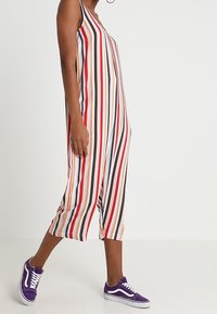 Une combinaison sans manches à rayures verticales rouges, noires, orange et blanches. Portée avec des baskets violettes, longueur cheville et coupe décontractée.