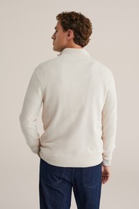 Cremfarbener Strickpullover mit strukturiertem Finish, gerippten Bündchen und Saum sowie einem hohen Kragen, kombiniert mit dunklen Jeanshosen.