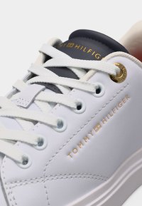 Κοντινό πλάνο λευκού αθλητικού παπουτσιού Tommy Hilfiger με λευκά κορδόνια, χρυσό μεταλλικό τρύπα και μπλε γλώσσα που φέρει το όνομα της μάρκας σε χρυσά γράμματα.