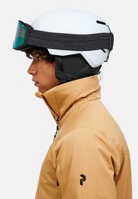 Casco da sci bianco con cinturino nero, abbinato a una giacca color marrone chiaro. Il casco ha fori di ventilazione e una visiera con sfumatura verde.