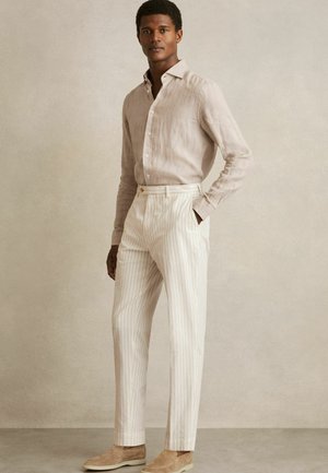 Cămașă din in beige deschis, pantaloni alb-crem cu dungi verticale și pantofi slip-on bej. Croială adaptată cu o textură netedă.