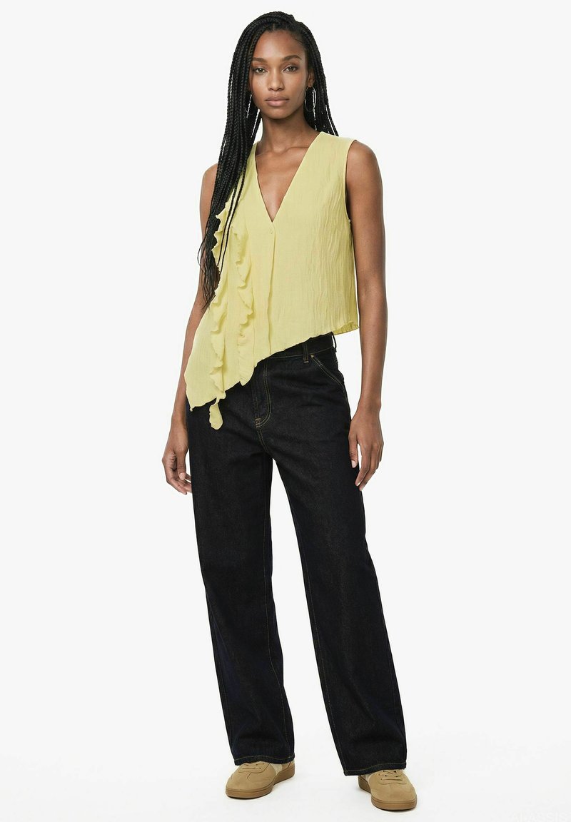 Femme aux longs cheveux tressés portant un top jaune sans manches à volants, un jean noir à larges jambes et des chaussures beiges, debout sur un fond blanc.