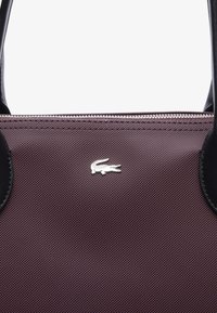 Sac à main en tissu bourgogne avec des accents en cuir noir, arborant un logo de crocodile argenté et une fermeture à glissière sur le bord supérieur.