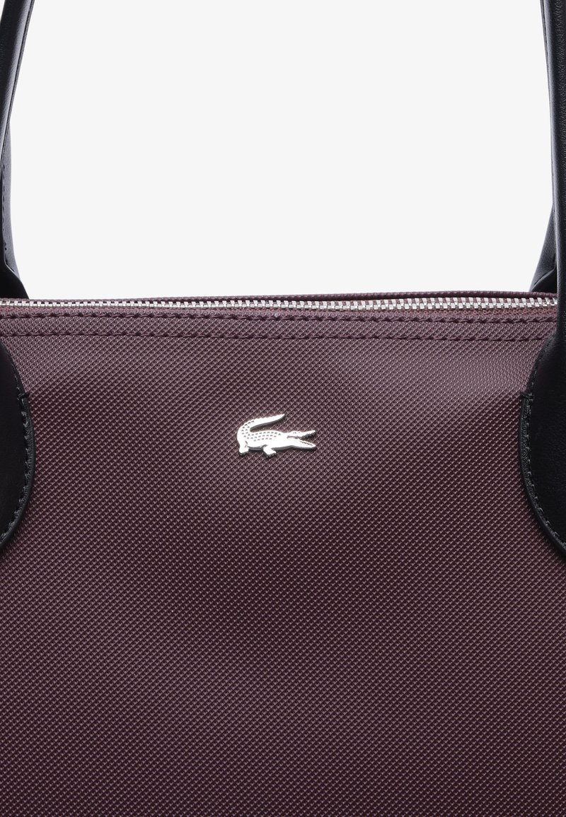 Burgundowa torba materiałowa z czarnymi skórzanymi akcentami, z logo w kolorze srebrnym w kształcie krokodyla i zamkiem błyskawicznym na górnej krawędzi.