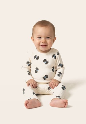Smiješeća beba sjedi u kremasto obojenom pidžamu s crnim otiscima pasa na jednobojnoj svijetloj pozadini.