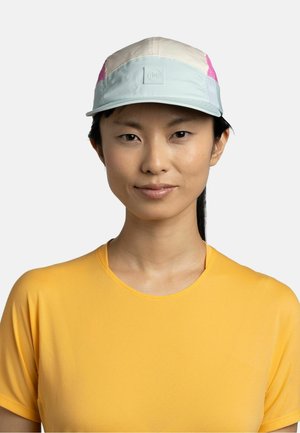 Frau mit glatten schwarzen Haaren, die eine pastelldfarbene Kappe und ein knallgelbes kurzärmeliges Shirt trägt und vor einem schlichten Hintergrund nach vorne schaut.