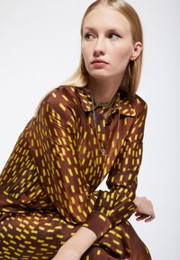 Vestito in satin marrone con un motivo rettangolare giallo, dotato di colletto e maniche lunghe. Gli accessori includono collane a strati e orecchini a cerchio.