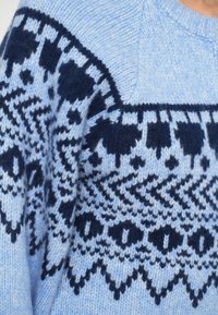 Modrá pletená sveter s geometrickým vzorom v tmavomodrej farbe, s mäkkou textúrou a okrúhlym výstrihom. Prvky zahŕňajú motívy v tvare zigzagu a bodiek.