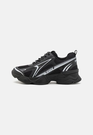 Chaussures de sport noires avec une tige en mesh, accents blancs, semelle épaisse et forme arrondie. Présentent des lacets et une boucle à l'arrière.