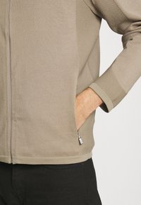 Béžová bunda na zip s texturovanými rukávy, žebrovanými manžetami, bočními kapsami a přiléhavým střihem, doplněná černými kalhotami.