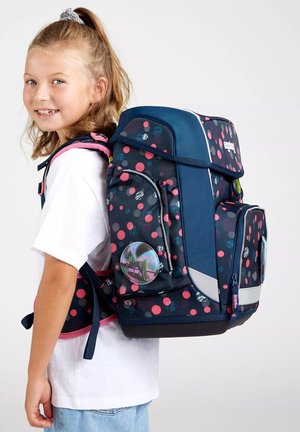 Marinefarbene Rucksack mit pinkem Punktmuster, mehreren Reißverschlussfächern, reflektierenden Akzenten und einem runden, blasenartigen Aufnäher an der Seite.