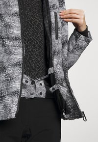 Pikowana kurtka w czarno-białym wzorze węża, z zasuwaną kieszenią i zapięciami na napy. Nosi ją na fakturowanej czarnej bluzce.