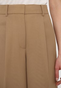 Pantalons beige ajustés avec une taille haute, des plis et une texture lisse. Passants de ceinture intacts, sans matériel visible ni détails supplémentaires.