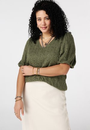 Femme aux cheveux noirs bouclés portant un pull texturé vert olive à manches courtes, une jupe crème et plusieurs bracelets en or et argent.