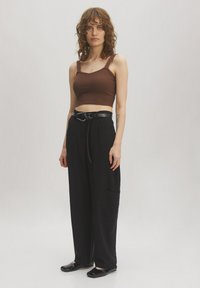 Braunes geripptes Crop-Top mit breiten Trägern, kombiniert mit locker sitzenden schwarzen Hosen mit einer Fronttasche und einem schwarzen Gürtel. Schwarze flache Schuhe.