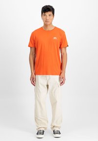 Oranges Baumwoll-T-Shirt mit Rundhalsausschnitt und kurzenÄrmeln, mit einem kleinen weißen Logo auf der Brust. Trägt man zu beigen Cargo-Hosen.