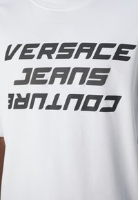 Bílá bavlněná tričko s výrazným černým textem, který diagonálně přes hrudník říká "VERSACE JEANS COUTURE".