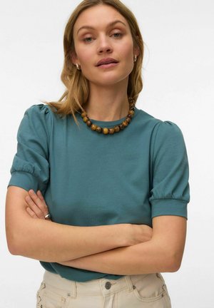 Woman with crossed arms wearing a teal puff-sleeve top, beige pants, a wooden bead necklace, and small hoop earrings against a plain background.