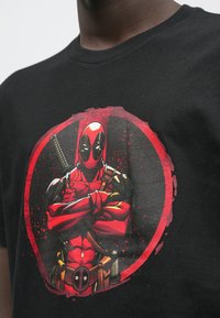 Mustast puuvillast T-särk, millel on värvikas graafika tegelasest punase ja musta kostüümiga, kes hoiab mõõka, punase ümmarguse taustaga.