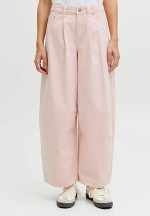 Person som har på seg lyserosa vide bukser og hvite joggesko, står med hendene avslappet langs sidene mot en ensfarget bakgrunn.