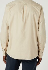 Camicia beige a maniche lunghe con yoke sulle spalle e dettaglio plissettato sul retro. Presenta polsini con bottoni, orlo arrotondato e tessuto strutturato.
