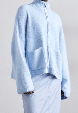 Weicher hellblauer flauschiger Reißverschluss-Pullover mit Fronttaschen und langen lockeren Ärmeln, getragen über einem hellblauen Rock.