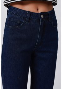 Pantalones vaqueros de mezclilla azul oscuro con cintura de altura media, cierre de botón plateado y diseño clásico de cinco bolsillos. Tela texturizada con un leve desgaste.