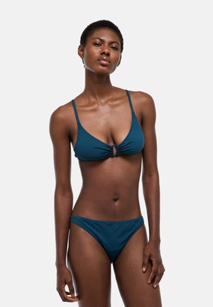 Femme mince portant un bikini deux pièces couleur sarcelle avec un détail œillet au niveau du haut, debout contre un fond gris clair uni.