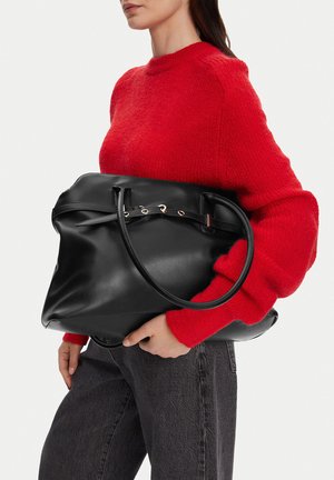 Mujer con un suéter rojo sosteniendo un gran bolso de cuero negro con un detalle de cinturón sobre un fondo liso.