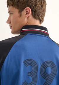 Blå och svart jacka med en ribbad krage med röda, vita och svarta ränder samt en framträdande svart, böjd "39" design på ryggen.