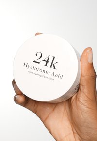 Conteneur rond et blanc étiqueté "Patch oculaire en hydrogel doré à l'acide hyaluronique 24k", tenu dans une main. Le couvercle présente un texte noir en gras.