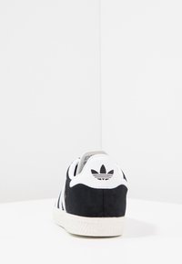 Svart och vit sneaker i mockamaterial, med tre vita ränder, en texturerad vit sula och en logotyp på hälfliken.