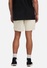 Light beige shorts with a simple design, featuring a folded hem and a small logo detail on the side. Worn with black high-top shoes.