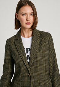 Blazer a quadri verdi e gialli con chiusura a pulsante singolo, colletto a scollatura, e tasche frontali, indossato sopra una t-shirt bianca con grafica.
