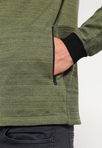 Olive green knit pullover with a zippered pocket, black cuff accents, and textured side panel. Features a smooth, casual design.