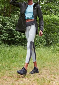 Zwarte waterdichte jas, lichtblauw aansluitend topje, grijze leggings met zwarte accenten en zwarte sportschoenen, tegen een groene achtergrond.