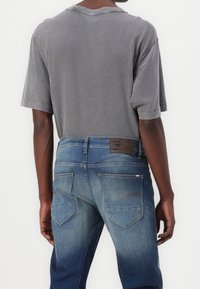 Šedé bavlněné tričko spárované s modrými džínami. Džíny mají vybledlý střih, hnědou koženou nášivku a dvojité zadní kapsy.